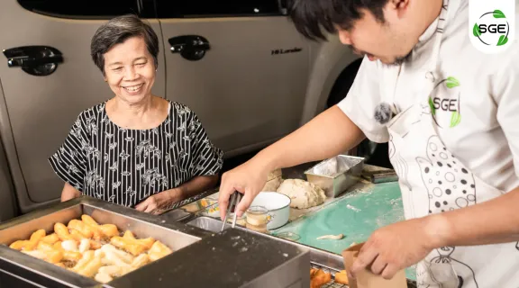 ผู้หญิงสูงวัย ใบหน้ากำลังยิ้มแย้ม มองผู้ชายทำกำลังทอดปาท่องโก๋ ในหม้อทอดน้ำน้ำมัน